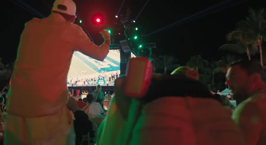 Guests watching the big game on a large outdoor screen at night during the Baha Mar Big Game Tailgate, with colorful stage lighting and palm trees.