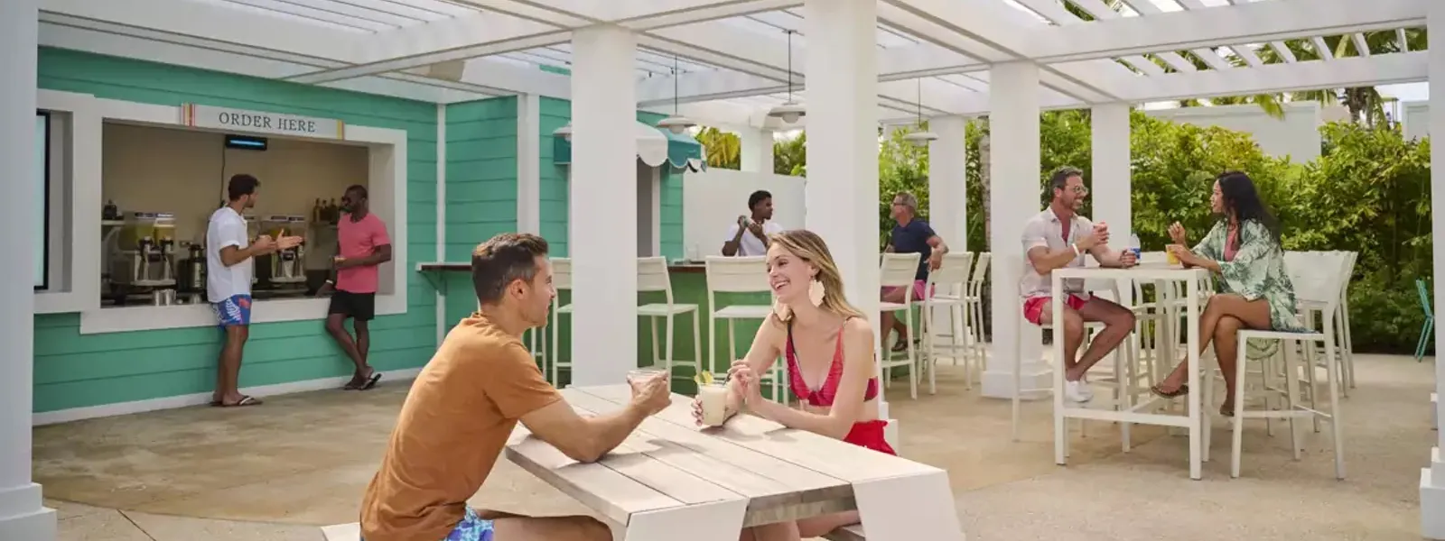 Guests enjoying drinks and food at The Studio café's outdoor terrace with turquoise order counter, white pergola seating, and tropical greenery.