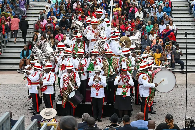 The Royal Bahamas Police Force marching band performing in white and red uniforms with festive holiday garlands, playing brass instruments and drums before a packed crowd in an outdoor amphitheater.