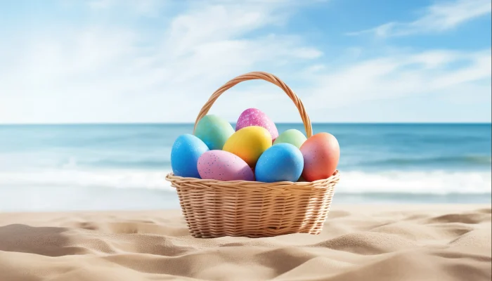 A wicker basket filled with colorful Easter eggs sitting on a sandy beach with a turquoise ocean and blue sky in the background.
